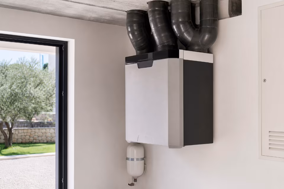 Bijeli zidni bojler s crnim ventilacijskim cijevima montiran je na bijeli zid pored prozora koji gleda na zeleni vrt.
