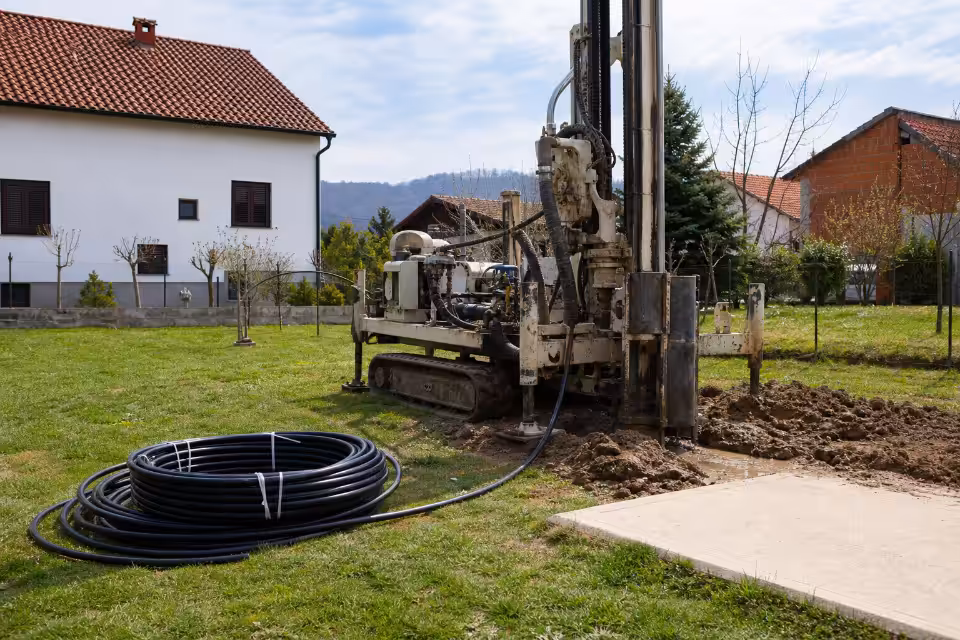 Buražna mašina na gusjenicama buši bunar u travnjaku uz kuću, uz kotur crne cijevi.