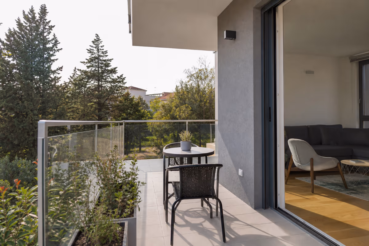 Balkon s staklenim ogradama, stolom i dvama stolicama, uz pogled na zelene drveće i otvorena vrata prema dnevnom boravku