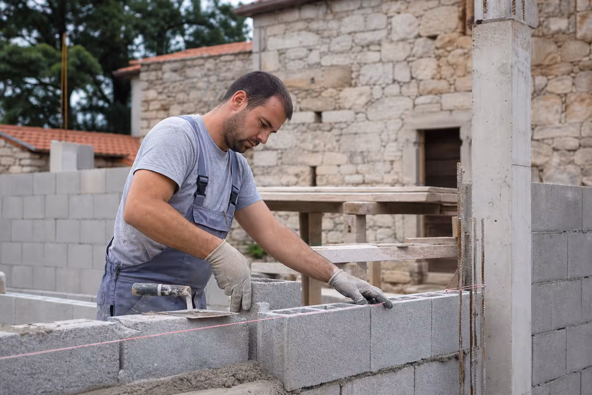 Zidar u radnom odijelu postavlja betonske blokove uz pomoć vodilice, ilustrirajući preciznost i profesionalnost ključne