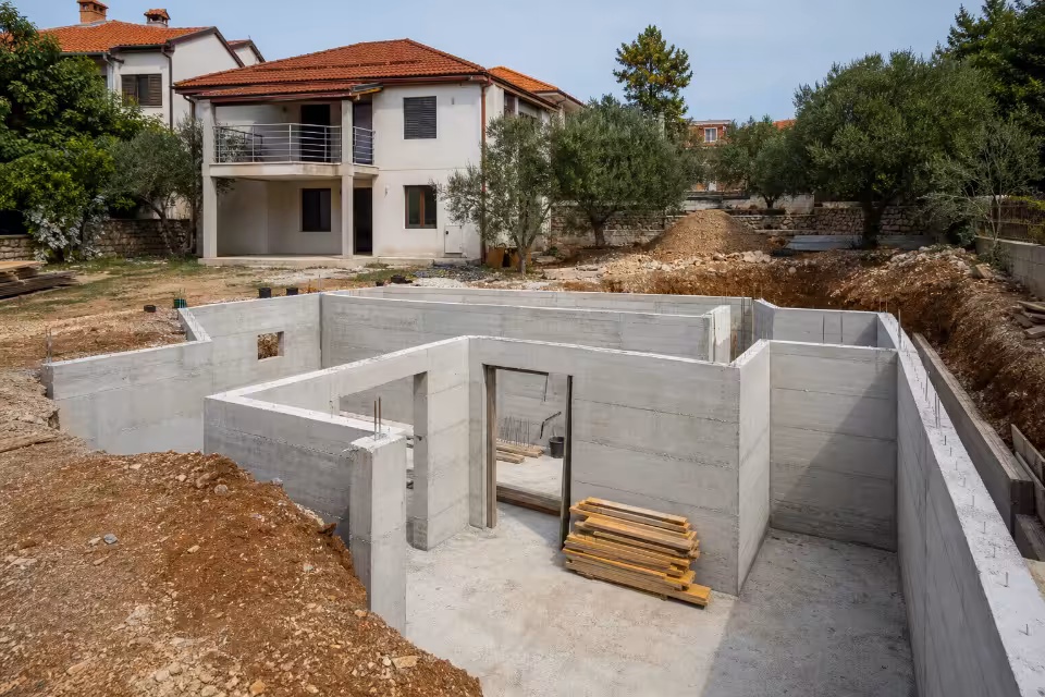 Betonski temelji i zidovi prizemlja nove kuće u izgradnji, okruženi zemljanim nasipima i građevinskim materijalom.