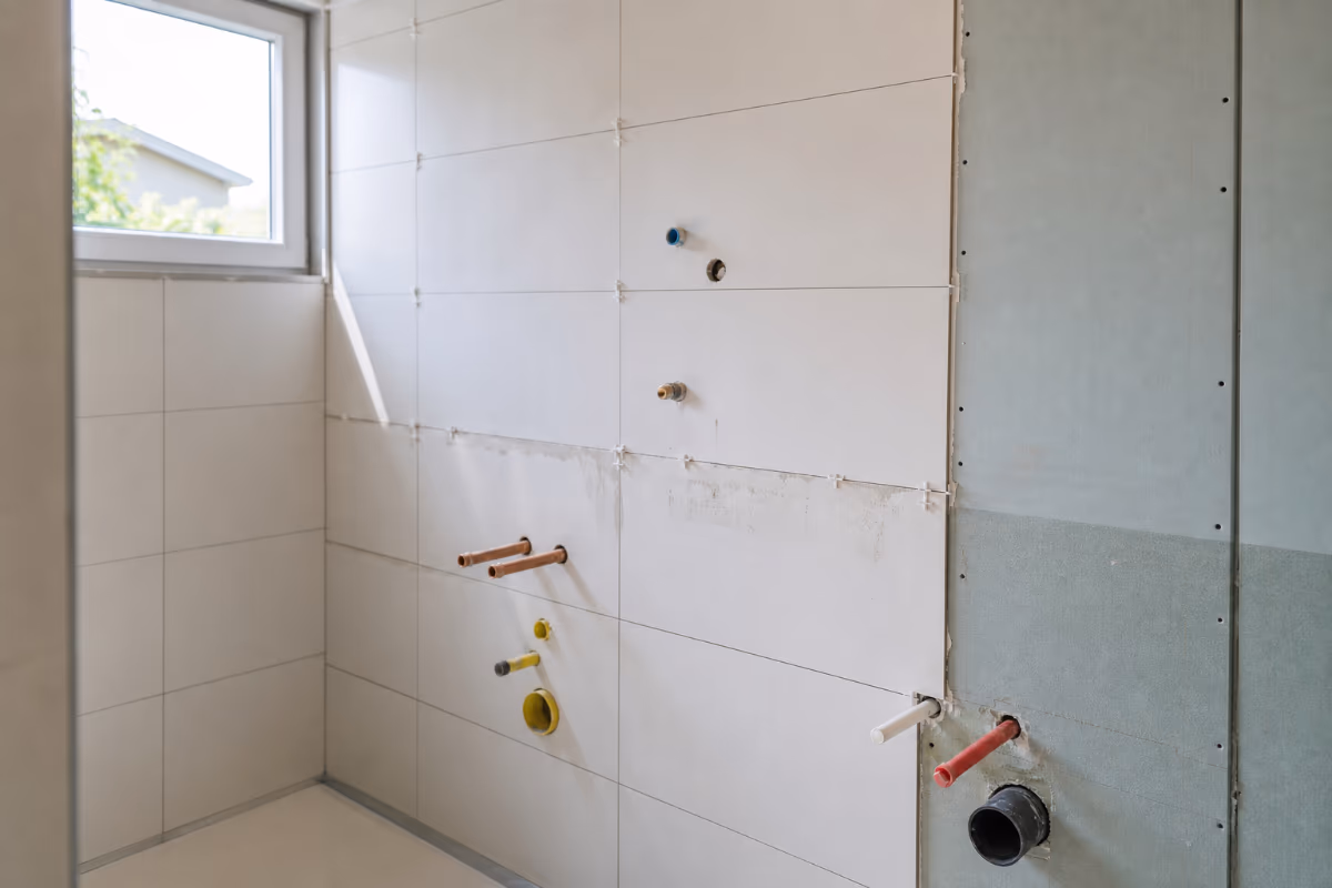 Kupaonica u gradnji s bijelim pločicama na zidu, izloženim vodovodnim cijevima i prozorom na lijevoj strani.