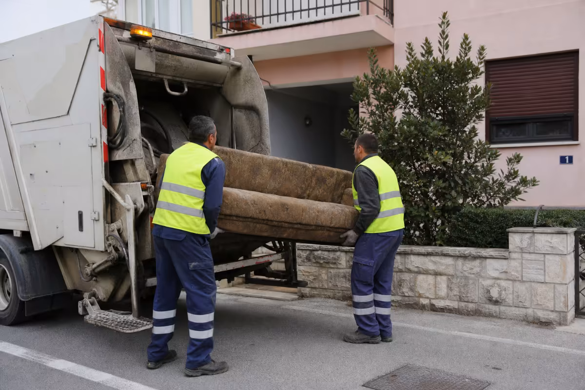 Radnici u reflektirajućim prslucima utovaruju staru sofu u kamion za odvoz glomaznog otpada.