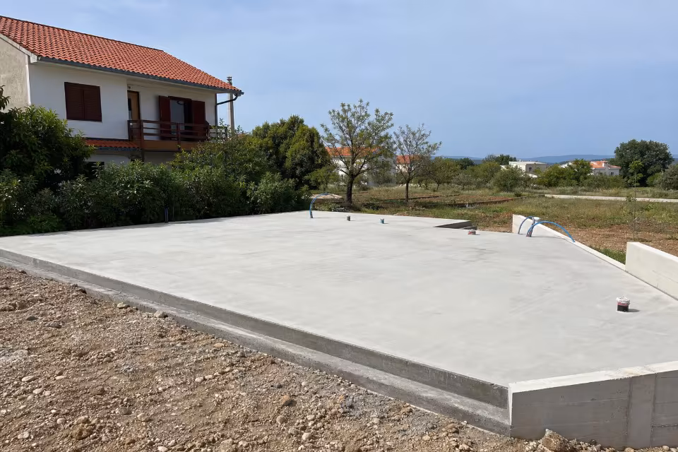 Sveže izlivena betonska temeljna ploča za gradnju kuće, s vidljivim instalacijama i okruženjem u izgradnji.