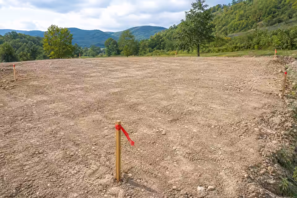 Prazan građevinski plac s drvenim kolcima i crvenim vrpcama na pozadini zelenih brežuljaka