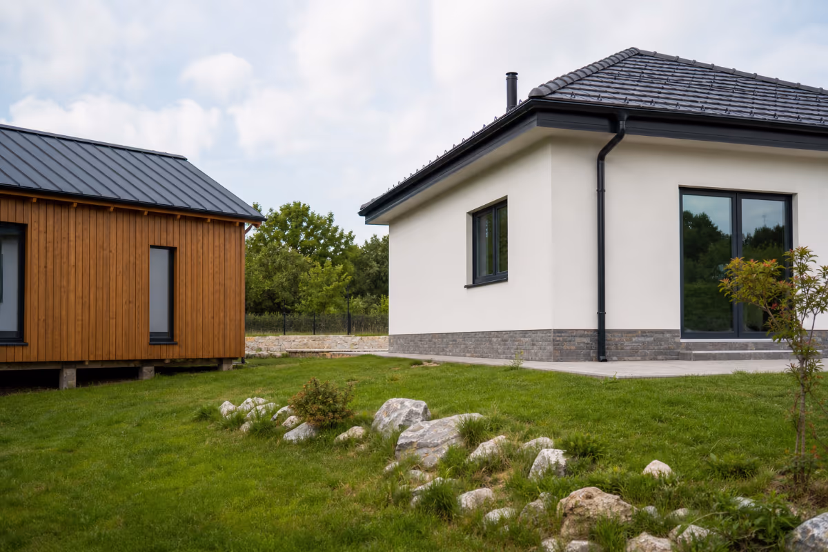 Moderna bijela kuća s crnim krovom i drveni objekt nalaze se na zelenom travnjaku s kamenjem, okruženi stablima pod obla