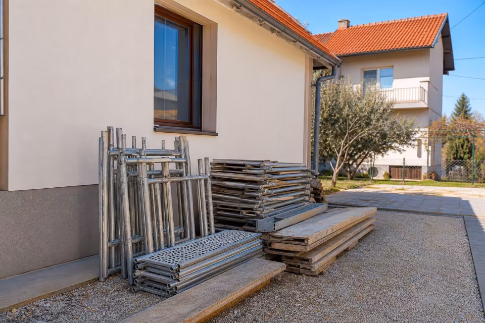 Na slici su vidljivi metalni elementi skela i drvene daske naslagane uz zid kuće na šljunkovitoj podlozi.