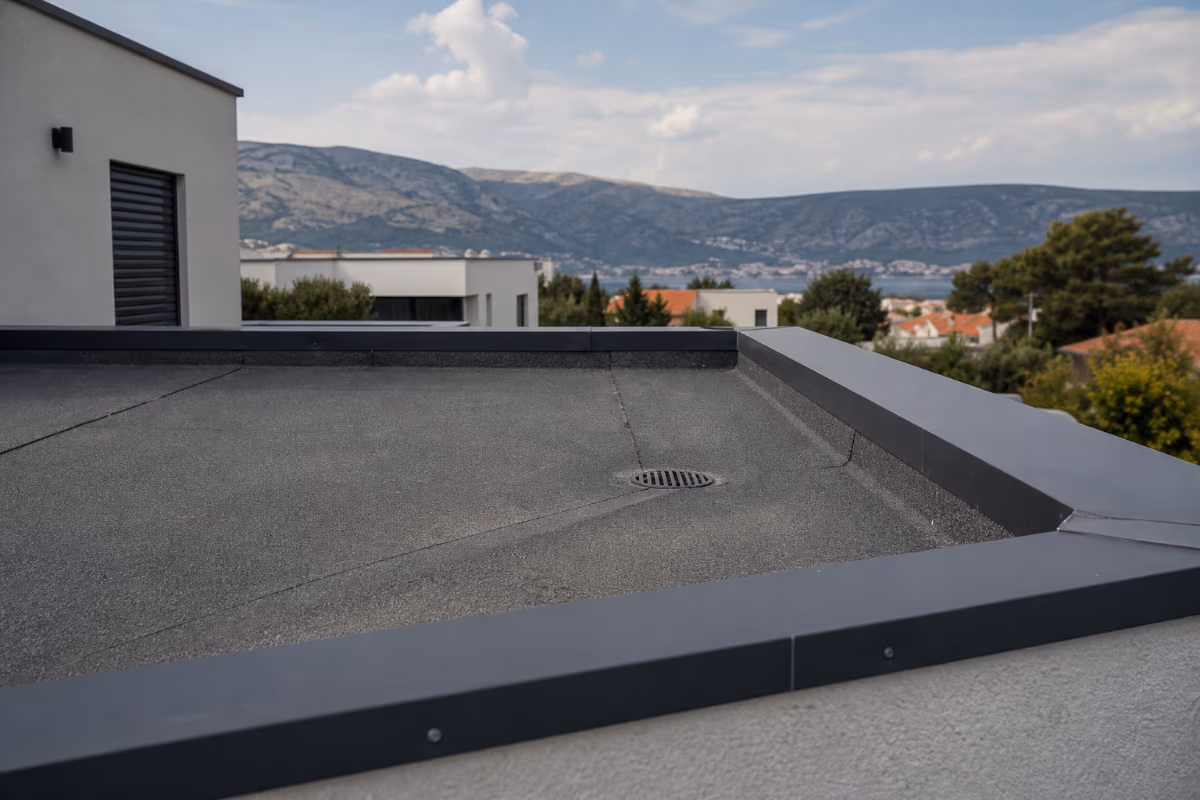Siva ravna krovna površina s metalnim rubovima i odvodnim otvorom, s pogledom na planine i naselje u pozadini.