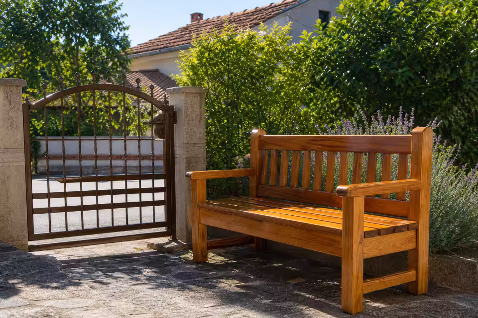 Sjajna drvena klupa stoji na kamenom dvorištu uz metalna vrata i zid od zelenog žbuna.