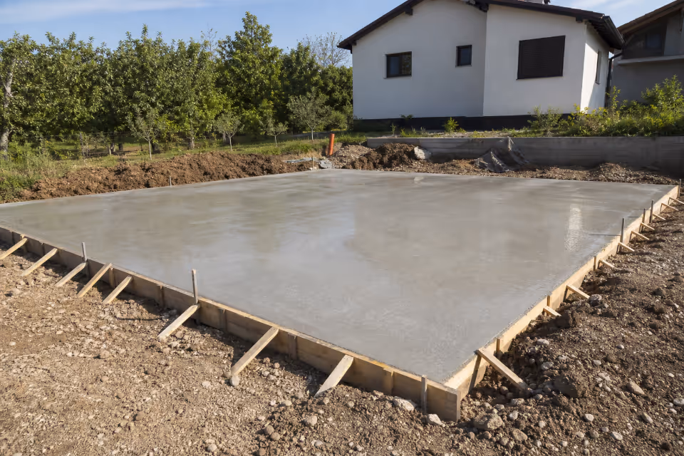Delavci v zaščitni opremi izlivajo beton za temeljno ploščo na gradbišču — podrobnost izvedbe