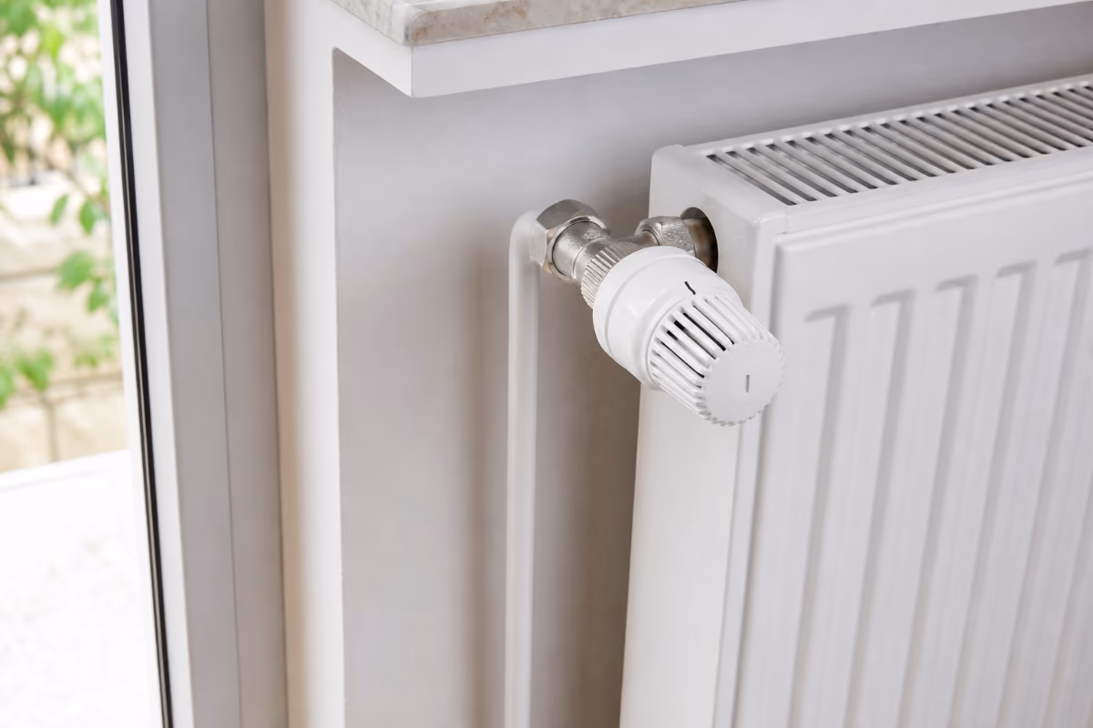 Bijeli radijator s termostatskim ventilom postavljen ispod prozora u svijetloj sobi.