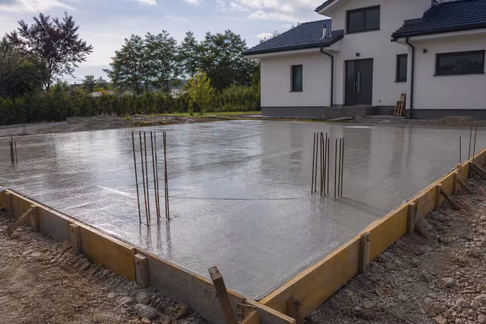 Delavci v zaščitni opremi izlivajo beton za temeljno ploščo na gradbišču — širši prikaz