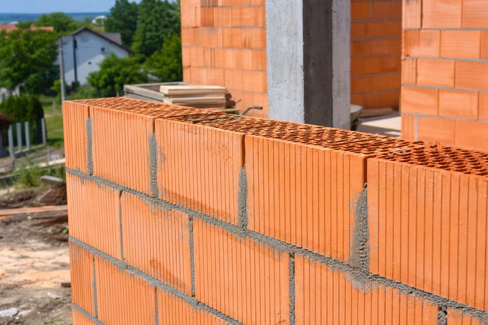 Građevinska radnja s novim zidom od crvene opeke i betonskim stupom, u pozadini se vide drveće i kuće.