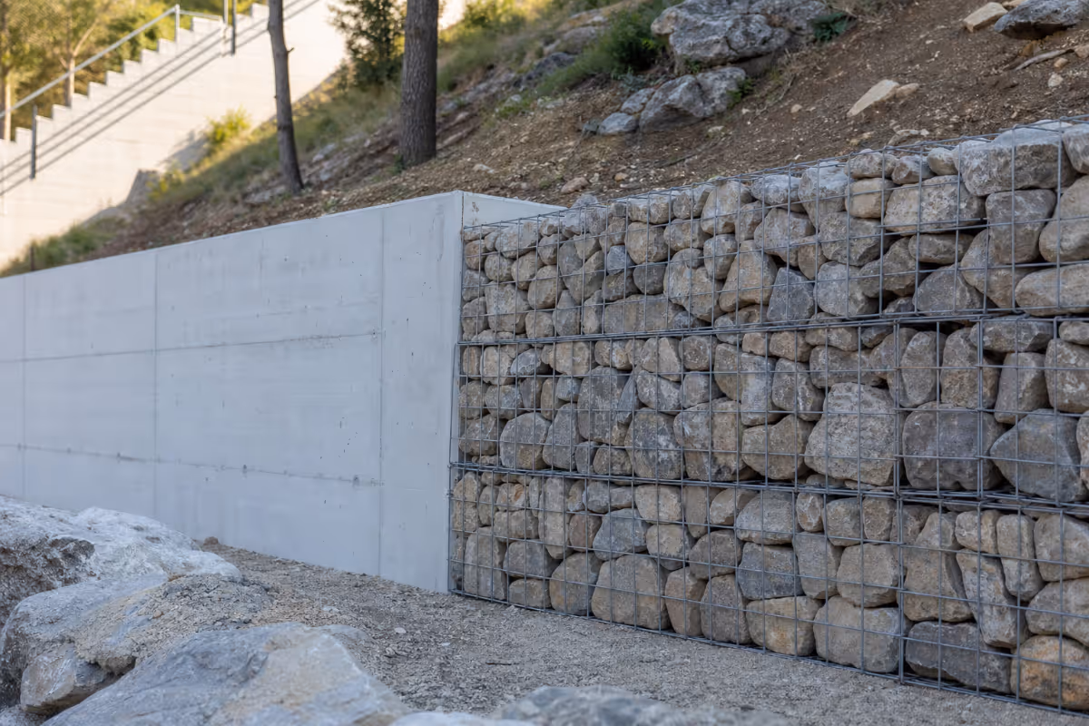 Detalj potpornog zida s bijelim betonskim panelima i gabionskim košarama ispunjenim kamenjem na obronku.