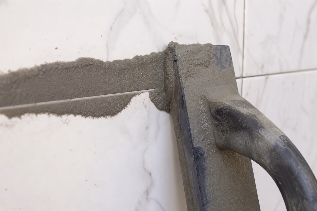 Detalj nanošenja cementne fug-mase metalnom lopaticom između bijelih pločica na zidu.