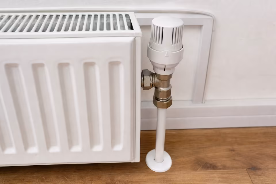 Bijeli radijator s termostatskim ventilom postavljen uz bijeli zid na drvenom podu.