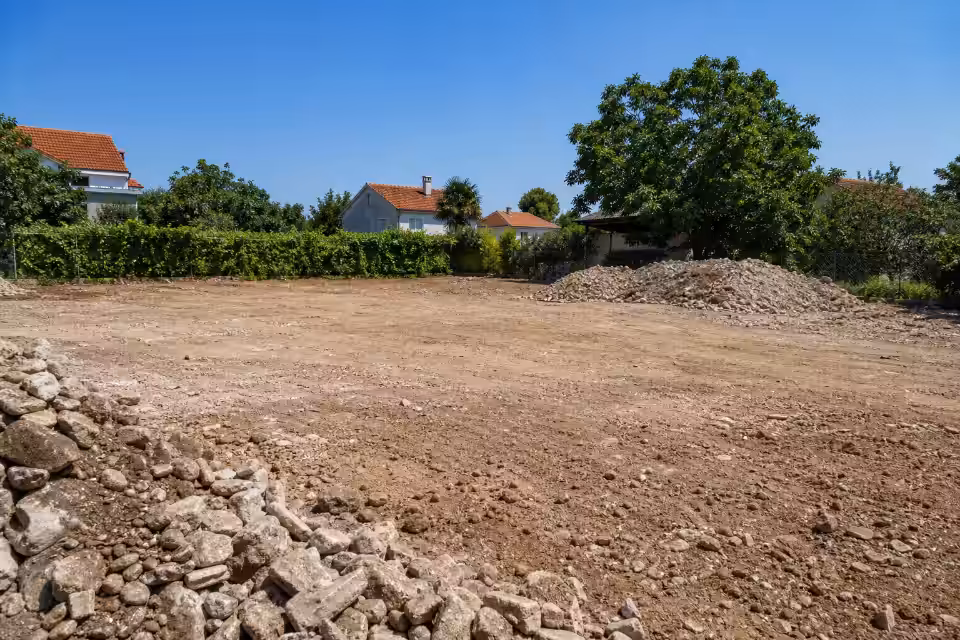 Poravnato tlo na lokaciji nakon rušenja kuće sa naslagama građevinskog otpada i zelenilom u pozadini