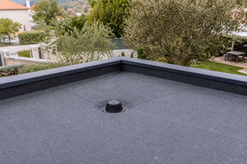 Siva ravna krovna ploha s crnim odvodnim otvorom u sredini i metalnim rubovima, okružena zelenilom i kućama u pozadini.