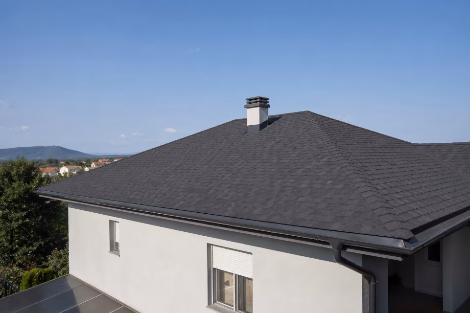 Krov s bitumenskim šindrama u tamnoj boji, uređen na modernoj kući s bijelim zidovima i dimnjakom, pod vedrim plavim nebom