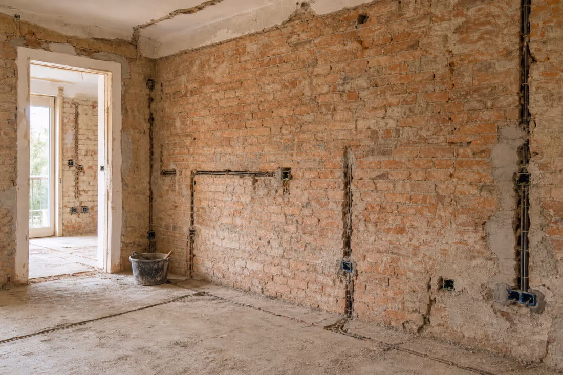 Prazna prostorija u fazi rušenja tijekom adaptacije, s ogoljenim ciglenim i betonskim zidovima, uklonjenim podom i usječenim kanalima za instalacije.
