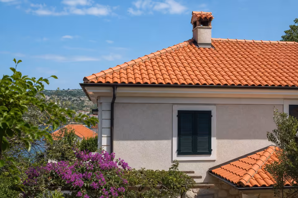 Kuća s narančastim crijepom i zelenim roletama, okružena cvjetnim grmljem i stablima uz plavo nebo.