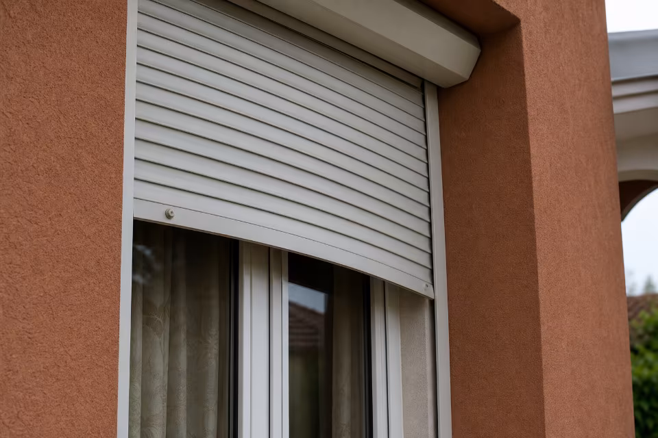 Bijela roleta djelomično spuštena pred prozorom na zgradi s narančastim zidom, kroz staklo se naziru zavjese.