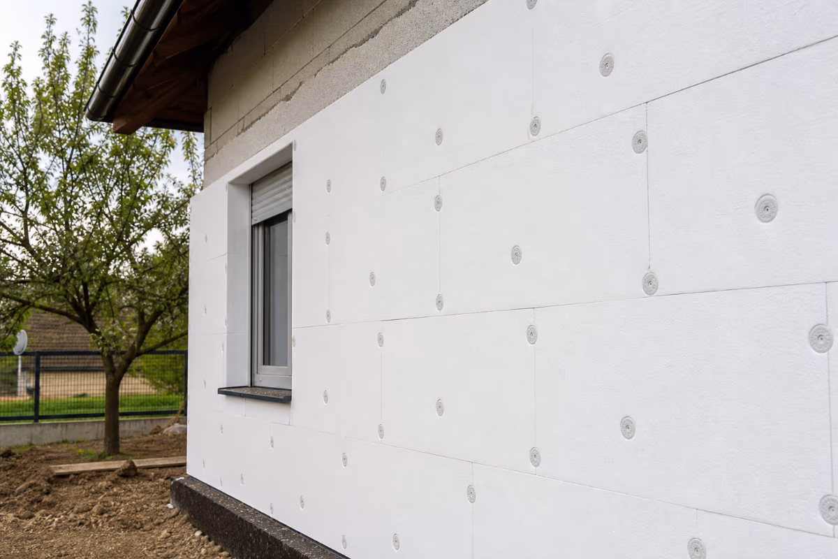 Vanjski zid kuće izoliran bijelim pločama s čepovima, u fazi gradnje, s prozorom i drvetom u pozadini.