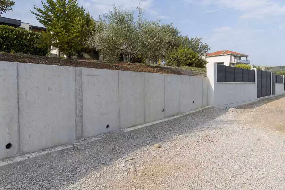 Siva betonska zidna ograda s crnim panelima proteže se uz šljunčanu cestu, a iza nje rastu stabla i kuće pod plavim nebo