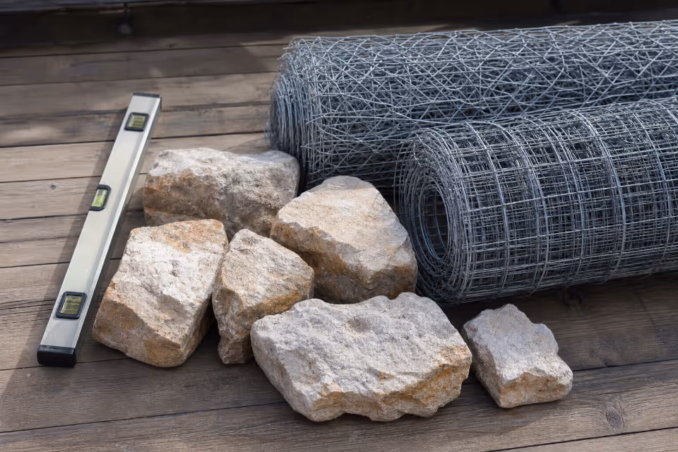 Gradivni materijali na drvenoj podlozi: prirodni kamen, rolovi čelične mreže za gabione i aluminijski vodokotlić.