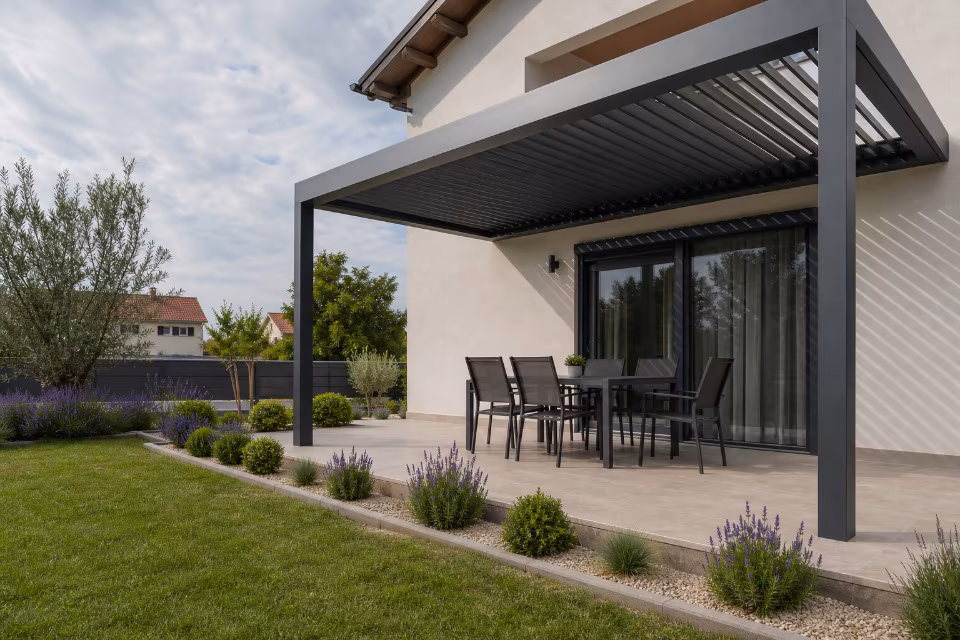 Črna kovinska pergola s poletno mizo in stolci na terasi moderne hiše ob urejenem vrtu z lavando in travnikom.