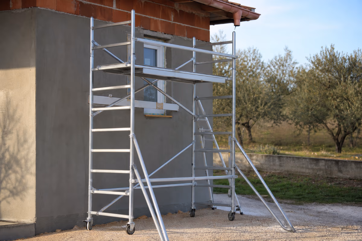 Aluminijska radna platforma postavljena uz zid kuće u izgradnji, s vidljivim prozorom i okolinom stabala masline.