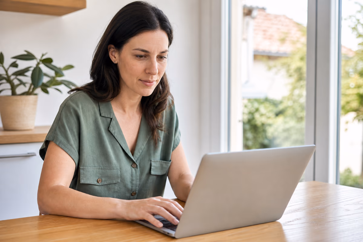 Žena u zelenoj bluzi sjedi za drvenim stolom i tipka na srebrnom laptopu uz prozor s pogledom na vrt.